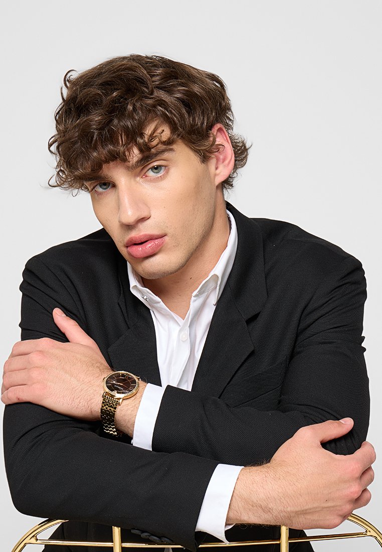 Zwarte blazer over een wit shirt, armen gekruist, met een gouden horloge met een bruine wijzerplaat. Krullend haar, neutrale achtergrond.