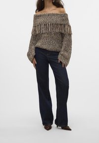 Sweter na ramiona, grubo dziergany, w brązowym i kremowym kolorze z ozdobnymi frędzlami, zestawiony z ciemnoniebieskimi, szerokimi spodniami jeansowymi oraz brązowymi butami z noskiem w szpic.