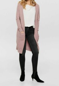 Cardigan lungo con apertura frontale in maglia rosa chiaro, dotato di polsini a coste e tasche, abbinato a una maglietta bianca e jeans skinny neri.