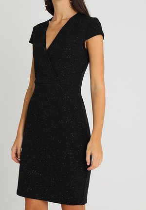 Femme portant une robe noire cintrée à manches courtes avec de fines taches blanches et un décolleté en V, debout devant un fond neutre uni.