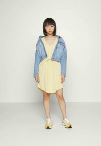 Vestido amarelo com cintura ajustável, combinado com uma jaqueta de ganga cropped. Modelo a usar ténis brancos e amarelos. Look simples e casual.