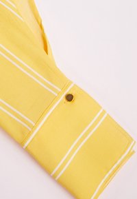 Polsino di camicia giallo con strisce bianche; texture liscia; presenta un bottone a scatto dorato e dettagli di cucitura.