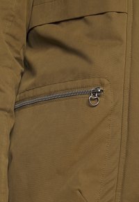 Olive green jacket fabric with a textured finish. Features a zippered pocket with a metal puller and visible stitching details.