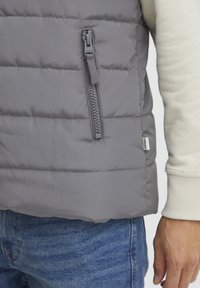 Grå puffervest med horisontalt quiltet mønster, med en sidelomme med glidelås og en liten merkeetikett nær kanten.