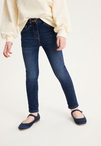 Jeans blu scuro a taglio slim, con chiusura a bottone e cerniera, abbinati a scarpe blu scuro flat con una fascia sopra. 