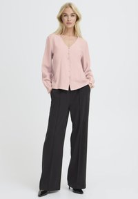 Blonde woman standing with hands in pockets wearing a pale pink long-sleeve buttoned blouse and black wide-leg trousers with heels.