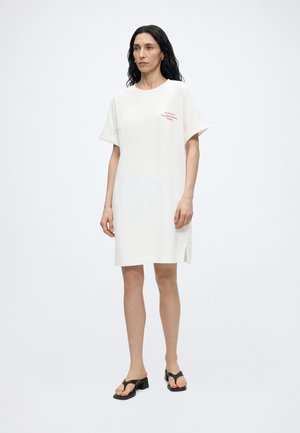 Γυναίκα με μακριά σκούρα μαλλιά που φοράει λευκό φόρεμα τύπου μπλουζάκι με κόκκινο κείμενο, στέκεται με μαύρα σανδάλια με τακούνι σε λευκό φόντο.