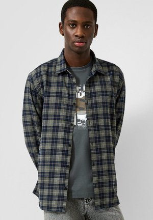 Jeune homme aux cheveux courts portant des boucles d'oreilles en anneaux dorés, un t-shirt graphique gris et une chemise à carreaux bleue et verte ouverte.