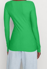 Kvinna som bär en åtsittande grön långärmad topp och ljusblå högmidjade byxor med vida ben, stående med ryggen vänd mot kameran.