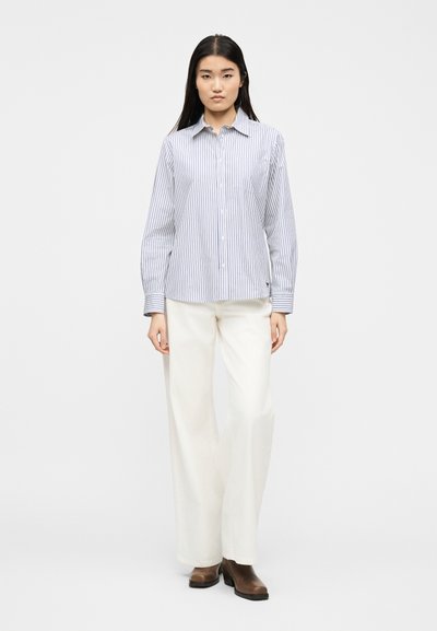 Γυναίκα που στέκεται, φοράει λευκό πουκάμισο με κάθετες μπλε ναυτικές ρίγες, λευκό παντελόνι με φαρδιές παντελόνες και καφέ μποτάκια αστραγάλου, με φόντο απλή επιφάνεια.