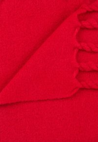 Coperta rossa in lana con una texture morbida, caratterizzata da un bordo a treccia e un angolo piegato, che mostra un colore vibrante e uniforme.