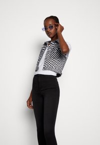 Top cropped em padrão xadrez preto e branco por cima de uma camisa branca, combinado com jeans ajustados pretos. O modelo usa óculos de sol roxos, acrescentando um toque moderno.
