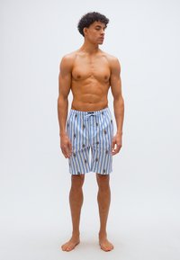 Άνδρας χωρίς πουκάμισο με σγουρά μαλλιά, που φορά μπλε και λευκό μαγιό με κάθετες ρίγες διακοσμημένο με μικρές εικόνες σκύλων, όρθιος ξυπόλητος σε λευκό φόντο.