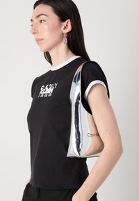 Calvin Klein silver metallic shoulder bag features a smooth texture, sleek design, and embossed logo, paired with a black t-shirt.
