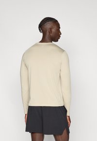 Μακρυμάνικη μπλούζα με στρογγυλή λαιμόκοψη σε ανοιχτό μπεζ ύφασμα, με λεία υφή και εφαρμοστή γραμμή· παρουσιάζεται με μαύρο σορτς που διαθέτει πλαϊνές σχισμές.