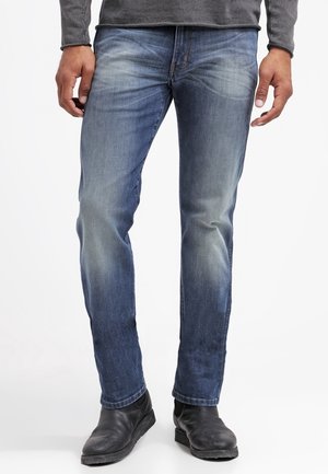 Mężczyzna ubrany w wyblakłe niebieskie dżinsy i czarne skórzane buty stoi z rękami swobodnie opuszczonymi wzdłuż ciała na jednolitym białym tle.