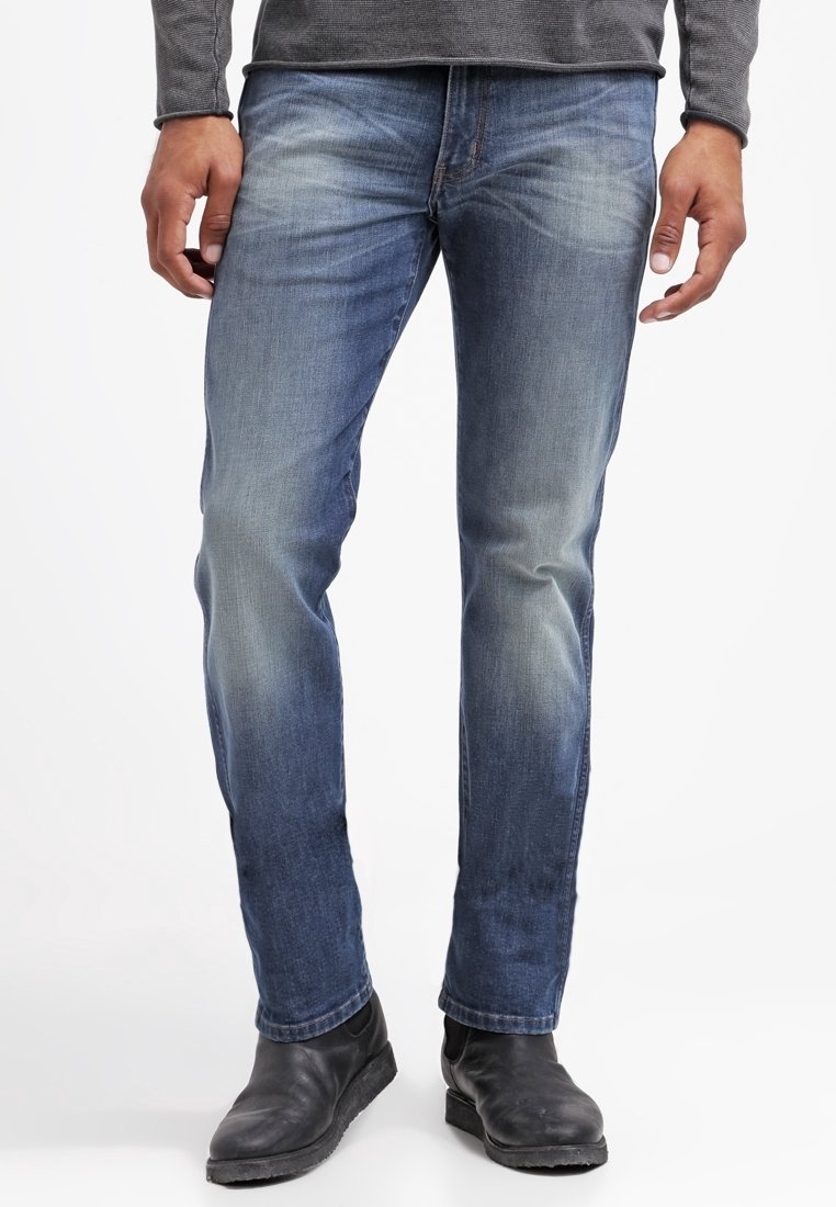 Mand iført falmet blå jeans og sorte læderstøvler, stående med hænder afslappet langs siden mod en enkel hvid baggrund.