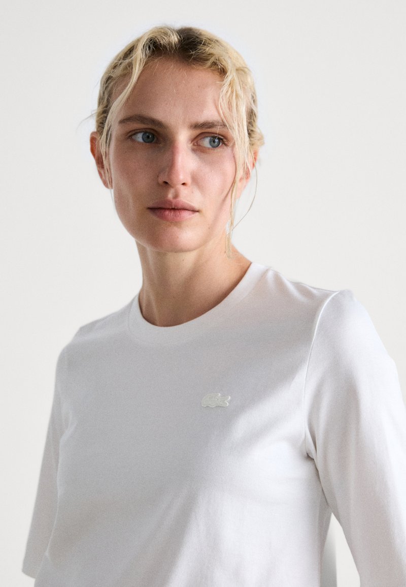 Biała bawełniana koszulka z okrągłym dekoltem i krótkimi rękawami, z małym haftowanym logo Lacoste na piersi. Miękka tekstura.