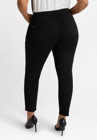 Svarta vadlångda byxor med elastisk midja, femfickorsdesign och slitna fållar, kombinerat med svarta högklackade skor.