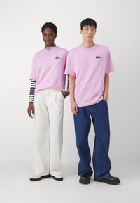 Due modelli in piedi, indossano camicie rosa a manica corta con un logo di coccodrillo verde; uno in pantaloni bianchi a gamba larga, l'altro in jeans blu.