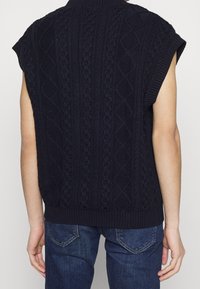 Gilet sans manches en tricot torsadé bleu marine porté par une personne en jean bleu foncé, vu de l'arrière.