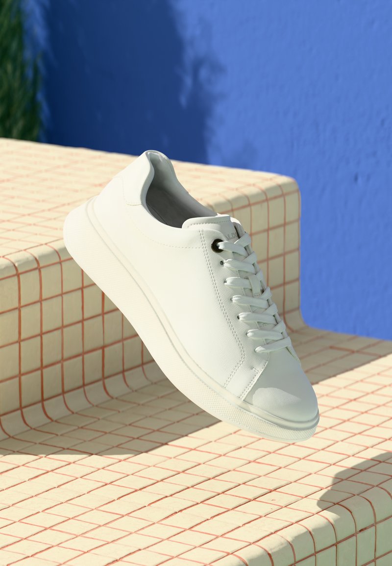 Baskets blanches en cuir lisse, avec un design bas, un bout rond et des lacets en tissu, posées sur une surface à motif en grille.