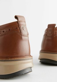 Chaussures en cuir brun avec une texture lisse, présentant un talon renforcé, une semelle intermédiaire beige contrastante et un petit logo en relief à l'arrière.
