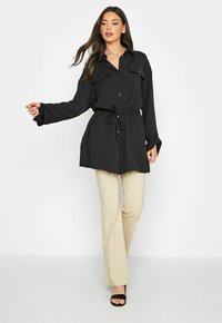 Zwarte blouse met lange mouwen, knoopsluiting, twee borstzakken, een trekkoord in de taille en een ontspannen pasvorm, gecombineerd met beige broek en zwarte hakken.