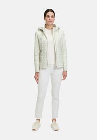 Giacca trapuntata di colore chiaro con cappuccio, dotata di zip frontale, tasche laterali e una texture liscia. Abbinata a una maglietta color crema e pantaloni.