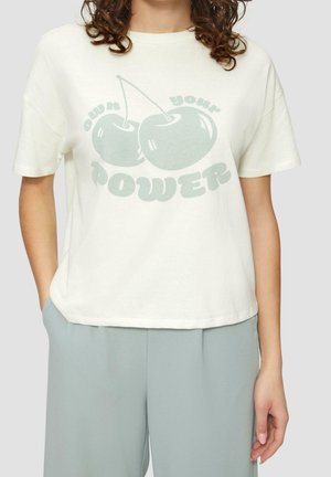 Kobieta nosząca białą koszulkę z dwoma jasnozielonymi wiśniami i napisem "own your power", połączoną z jasnoniebieskimi spodniami.