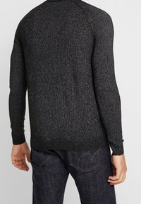 Pull noir en maille avec un motif texturé, poignets et ourlet côtelés, porté par une personne vêtue d'un jean foncé, vue de dos.