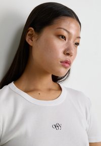 T-shirt bianco a costine con scollatura rotonda e un piccolo logo nero sul petto. Texture liscia, design aderente e maniche corte.