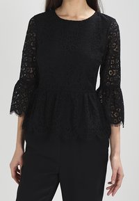 Top peplum de encaje negro con mangas acampanadas de tres cuartos, que presenta intrincados diseños recortados y un cuello redondo. Tejido texturizado.