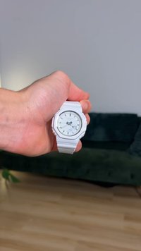 Λευκό ρολόι χειρός με στρογγυλό καντράν, μινιμαλιστικό καντράν, ασημένιες λεπτομέρειες και υφασμένο λουράκι, κρατημένο στο χέρι. Το φόντο παρουσιάζει έναν πράσινο καναπέ.