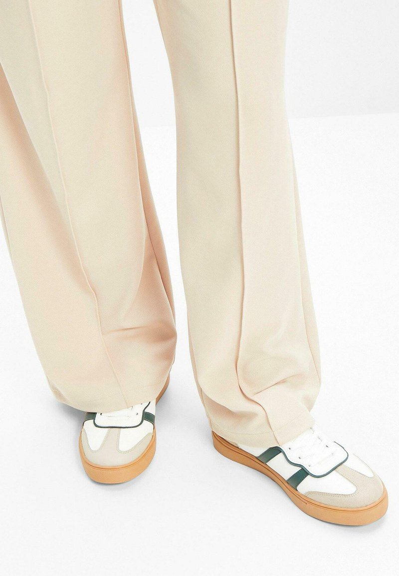 Scarpe da ginnastica bianche e grigie con una striscia verde, dotate di una tomaia testurizzata e una suola in gomma, abbinate a pantaloni beige larghi.