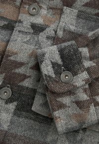 Ullskjorta med ett grått och brunt geometriskt mönster, med en bröstficka och en detalj med metalltryckknapp. Texturerad, varm tyg.