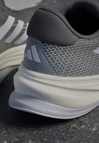 Baskets de sport grises avec une tige en maille texturée, présentant des rayures blanches et un talon rembourré doux. La semelle est gris clair avec des motifs subtils.