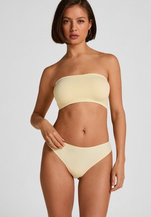 Frau mit kurzen braunen Haaren, die ein hellgelbes trägerloses Bandeau-Oberteil und passende Bikinihöschen vor einem einfarbigen Hintergrund trägt.