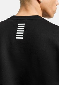 Felpa nera con strisce orizzontali bianche sulla parte superiore della schiena. La texture appare liscia e ha un colletto a girocollo.