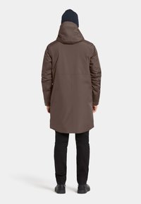 Ruskea eristysvuorattu takki hupulla, pitkä leikkaus ja olkasaumat. Yhdistetty mustiin housuihin ja mustiin kenkiin kerroksellisen ilmeen luomiseksi.