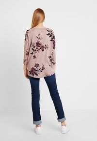 Helles rosa langärmeliges Oberteil mit floralem Muster in Lila und Schwarz, kombiniert mit dunkelblauen schmalen Jeans, mit aufgerollten Bündchen und weißen Schuhen.