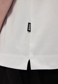 Biele bavlnené tričko s malou čiernou etikietou "BOSS" na leme. Tričko má bočný rozparok na spodnej časti pre zvýraznenie detailu.