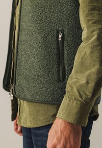 Green fleece vest with textured fabric, zippered pocket, and contrasting green button-up shirt underneath, paired with denim jeans.