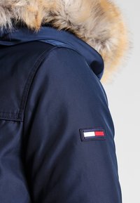 Mörkblå vinterjacka med axelparti och fleecefodrad huva samt rektangulär lappar med horisontella vita, röda och blå ränder.