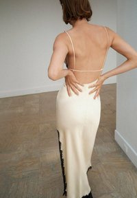 Une robe nuisette ajustée de couleur crème avec des fines bretelles, comportant des accents en dentelle noire le long de l'ourlet. Le dos est ouvert avec une texture lisse et élégante.