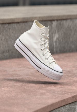Witte hoge sneaker met een zwarte streep op de zool, veters gestrikt, die lijkt te zweven boven een stenen oppervlak en een richel op de achtergrond.