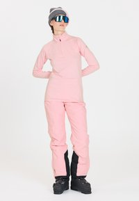 Rosa technische Langarmoberteil mit einem Viertel-Reißverschluss, kombiniert mit rosa Skihosen mit schwarzen Akzenten. Das Model trägt eine graue Mütze und verspiegelte Sonnenbrillen.