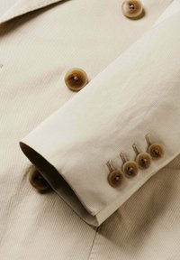 Manche de veste beige avec quatre boutons marron cousus en ligne près du poignet et deux boutons assortis sur le corps.