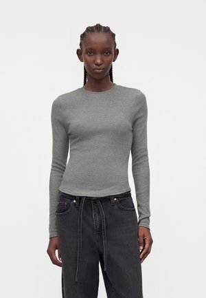 Γυναίκα με πλεξούδες που φοράει στενό γκρι μακρυμάνικο πουκάμισο και χαλαρά μαύρα τζιν με ζώνη, στέκεται μπροστά σε λευκό φόντο.