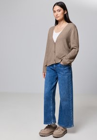 Beige kofta med v-ringning och knappar, bärs över en vit topp, kombinerad med vida blå jeans och ljusbruna skor. Slät textur.